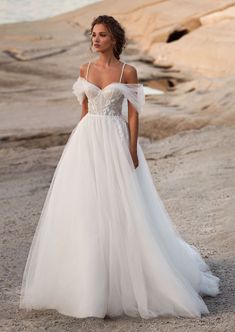 a woman in a white wedding dress standing on the beach with her hands behind her back