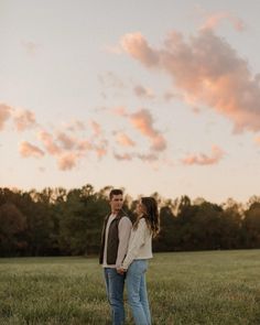 a man and woman standing in the middle of a field at sunset with clouds overhead