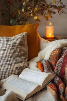 an open book sitting on top of a bed next to pillows and a lit candle