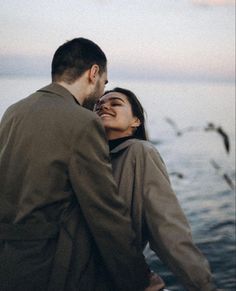 a man and woman standing next to each other near the water with seagulls flying overhead