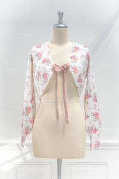 A cute and unique cardigan, the vintage-inspired Coquette bolero-style sweater features long sleeves, a lovely pink rose print, and a blush pink bow-style ribbon closure. . Details: S: Bust 38", Length 16" M: Bust 40", Length 17" L: Bust 42", Length 18" Length Measured from Shoulder Material: 50% Nylon, 20% Wool, 15% Polyester, 15% Acrylic Hand Wash Cold/Flat Dry/Dry Clean Imported Pink Long Sleeve Shrug For Spring, Long Sleeve Pink Shrug For Spring, Feminine Luxury Pink Cardigan, Feminine Pink Long Sleeve Cardigan, Bow Cardigan, Vintage Pink Winter Cardigan, Pink Bow Cardigan, Unique Cardigan, Bolero Cardigan