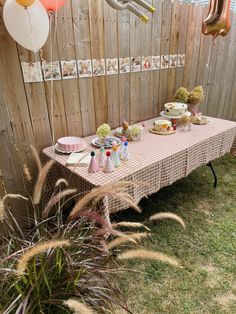 a table set up for a birthday party with balloons and cake on it in front of a wooden fence