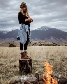 a woman standing on top of a tree stump next to a fire in a field