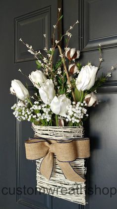 white flowers are in a basket hanging on the front door with burlap ribbon