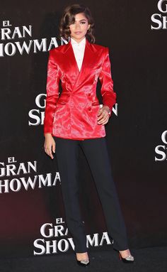 a woman in a red jacket and black pants posing for the camera at an event