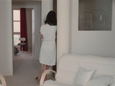 a woman in a white dress standing next to a bed and looking at the wall