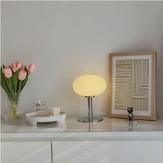 a vase with flowers and a lamp on a white shelf next to a framed photograph