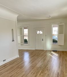 an empty living room with hard wood flooring and white walls, two windows on either side of the door