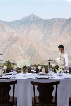 a man standing next to a table covered in white cloths and place settings with mountains in the background