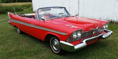 an old red car is parked in the grass near a white building and a fence