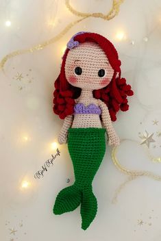 a crocheted little mermaid doll laying on top of a white surface next to christmas lights