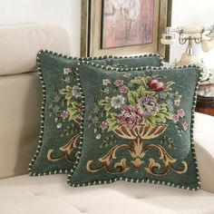 two decorative pillows sitting on top of a white couch next to a framed painting and lamp