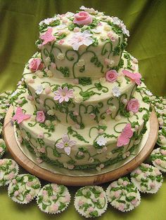 a three tiered cake with pink and green flowers on the top is surrounded by cupcakes