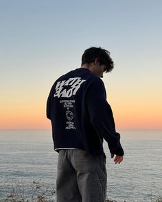 a man standing on top of a cliff next to the ocean at sunset with his hands in his pockets