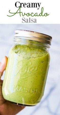 a hand holding a mason jar filled with creamy avocado salsa and text overlay that reads, creamy avocado salsa