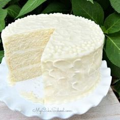 a white cake sitting on top of a white plate next to green leaves in the background