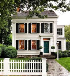 a white house with black shutters on the front and green door, surrounded by greenery