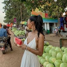 a woman in a white dress holding a piece of watermelon at an outdoor market