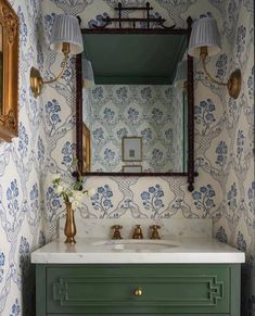 a bathroom vanity with green drawers and gold fixtures on the mirror above it is decorated in floral wallpaper