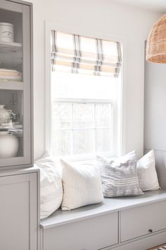 a white window seat with pillows on it in front of a cabinet and light fixture