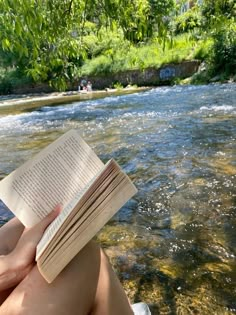 a person is reading a book while sitting on the edge of a river and holding it in their hands