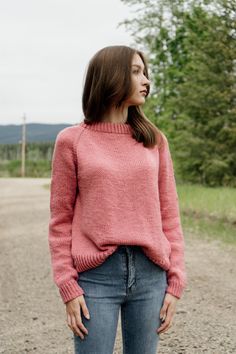 a woman standing in the middle of a dirt road wearing a pink sweater and jeans