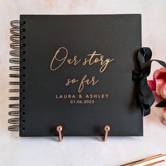 a black notebook with gold foil lettering on the cover and two pink flowers next to it