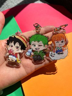 a hand holding four anime key chains on top of a colorful surface with other items in the background