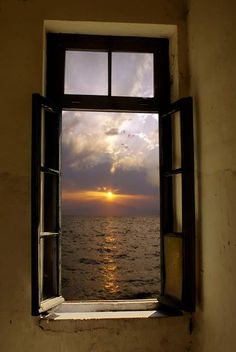 an open window with the sun setting over the ocean in front of it on a cloudy day