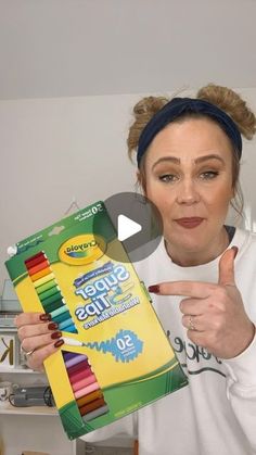 a woman holding up a box of crayons and pointing to the camera with her finger