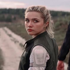 a woman with blonde hair wearing a green vest and looking at the camera while standing next to a man