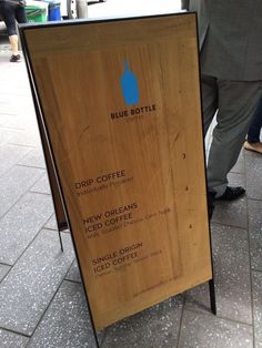 a wooden sign sitting on the side of a sidewalk next to a sidewalk with people walking by