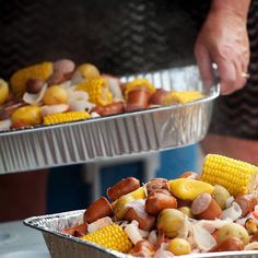 two metal pans filled with corn on the cob and sausage covered hot dogs