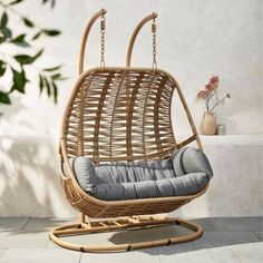 a rattan swing chair with grey cushions in front of a white wall and potted plant