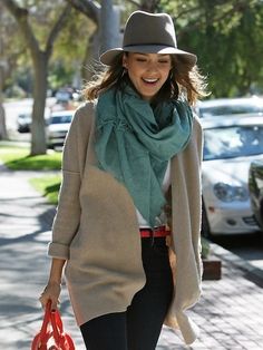 a woman walking down the street wearing a hat, scarf and sweater with her handbag