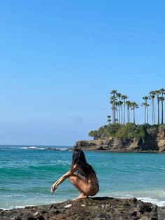 a woman sitting on top of a rock next to the ocean with palm trees in the background