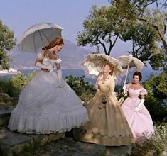 three women dressed in dresses and holding umbrellas on a hill overlooking the water with trees behind them