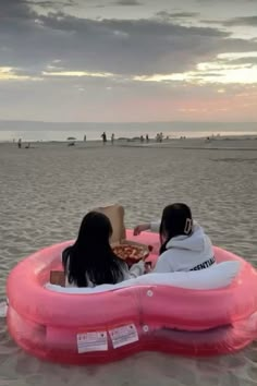 two women sitting in an inflatable boat on the beach at sunset, eating pizza