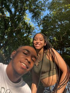 a man standing next to a woman with long braids on her head and trees in the background