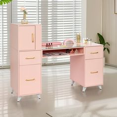 a pink desk with gold handles and drawers in front of a large window, next to a potted plant