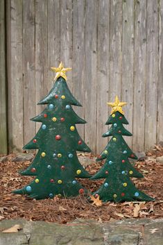 two green christmas trees sitting on top of leaves in front of a wooden fence with yellow stars