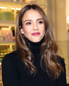 a woman with long brown hair wearing a black turtle neck sweater and pink lipstick looks at the camera