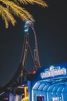 a roller coaster at night with lights on it's side and palm trees in the foreground