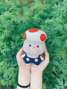 a hand holding a small crocheted doll in front of green plants