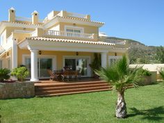 a large yellow house sitting in the middle of a lush green field next to a palm tree