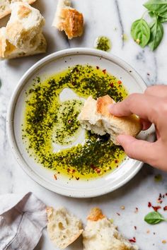 someone is dipping pesto on bread in a white bowl next to garlic bread and basil