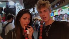 two young people standing next to each other eating food at an outdoor market place in the city