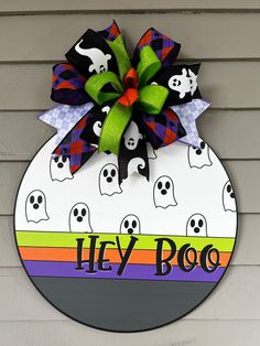 a colorful door hanger with ghost faces and green bow on it that says hey boo