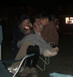 three people sitting in lawn chairs at night, one holding a lit candle and the other looking at her phone