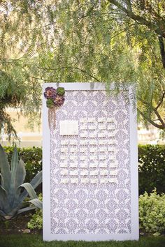 a white and purple wedding ceremony sign in front of a tree with succulents